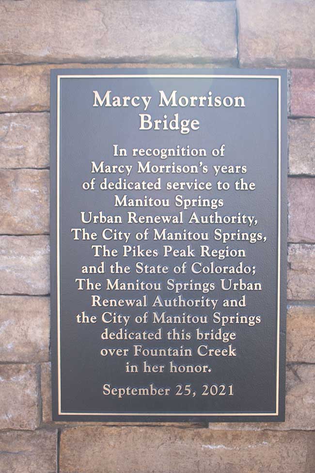This photo shows the Sign on the Marcy Morrison Bridge, which reads: In recognition of Marcy Morrison's years of dedicated service to the Manitou Springs Urban Renewal Authority, The City of Manitou Springs, The Pikes Peak Region and the State of Colorado; The Manitou Springs Urban Renewal Authority and the City of Manitou Springs dedicated this bridge over Fountain Creek in her honor. September 25, 2021. Success Stories from the Manitou Springs Urban Renewal Authority. This photo shows the Sign on the Marcy Morrison Bridge, which reads: In recognition of Marcy Morrison's years of dedicated service to the Manitou Springs Urban Renewal Authority, The City of Manitou Springs, The Pikes Peak Region and the State of Colorado; The Manitou Springs Urban Renewal Authority and the City of Manitou Springs dedicated this bridge over Fountain Creek in her honor. September 25, 2021. Success Stories from the Manitou Springs Urban Renewal Authority.