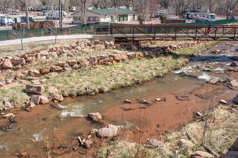 This photo shows the Creek Walk Trail with a bridge spanning over the creek and person crossing the new bridge. Success Stories from the Manitou Springs Urban Renewal Authority. This photo shows the Creek Walk Trail with a bridge spanning over the creek and person crossing the new bridge. Success Stories from the Manitou Springs Urban Renewal Authority.